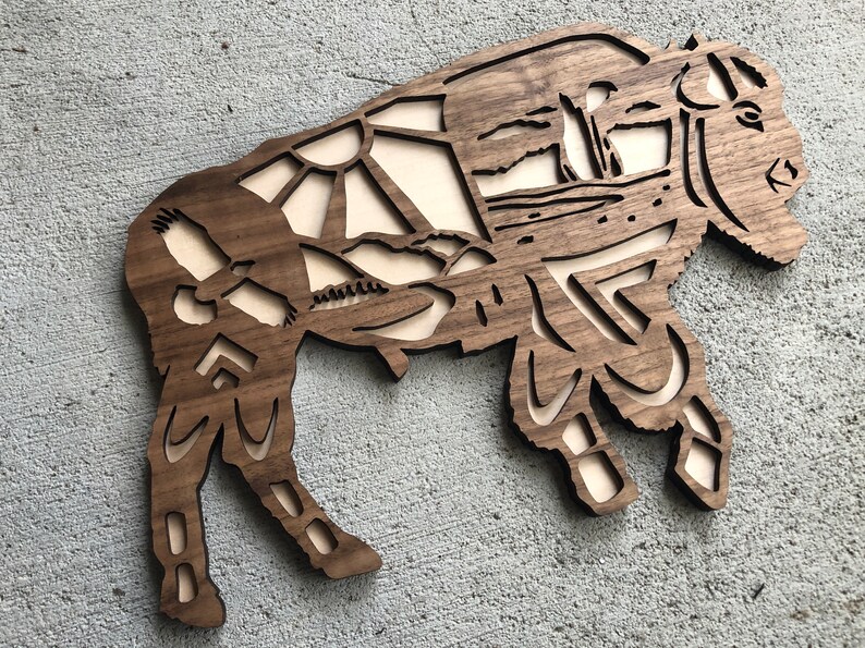 May include: A wooden wall art sculpture of a bison with intricate geometric patterns and a stylized landscape design. The bison is brown and the background is a light wood color.