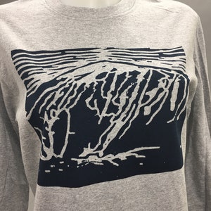 May include: A gray long-sleeve shirt featuring a dark blue graphic design. The graphic depicts a stylized mountain range with horizontal lines above. The shirt is displayed on a mannequin.