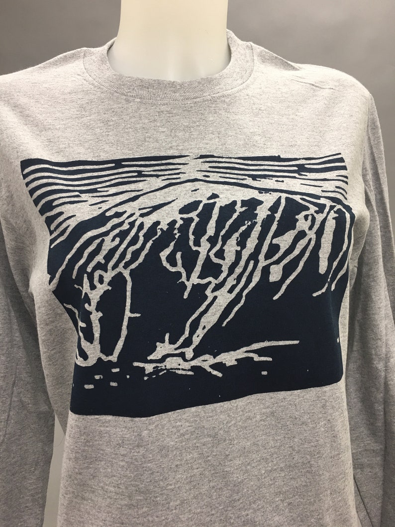May include: A gray long-sleeve shirt featuring a dark blue graphic design. The graphic depicts a stylized mountain range with horizontal lines above. The shirt is displayed on a mannequin.