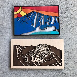 May include: Two wooden wall art pieces depicting a mountain range with ski runs. The top piece is painted in a blue, white, and orange color scheme with a sun design. The bottom piece is a dark brown wood with a white cutout of the mountain range.