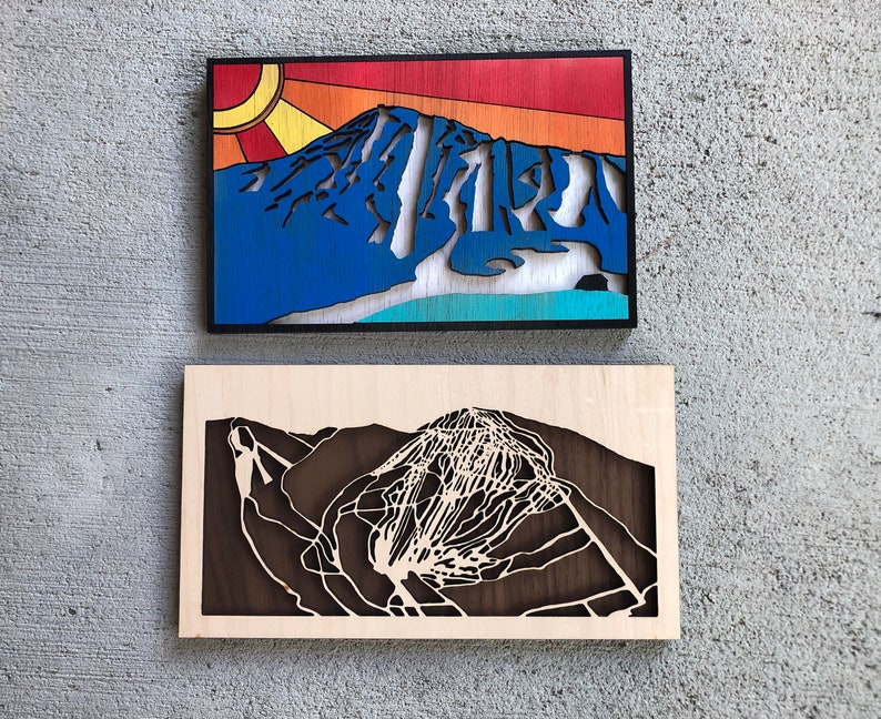 May include: Two wooden wall art pieces depicting a mountain range with ski runs. The top piece is painted in a blue, white, and orange color scheme with a sun design. The bottom piece is a dark brown wood with a white cutout of the mountain range.