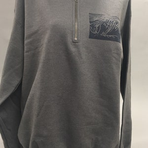 May include: A charcoal gray sweatshirt with a quarter-zip neckline and a small, dark blue square patch on the chest. The patch features a stylized, abstract design. The long-sleeved top is displayed on a mannequin.