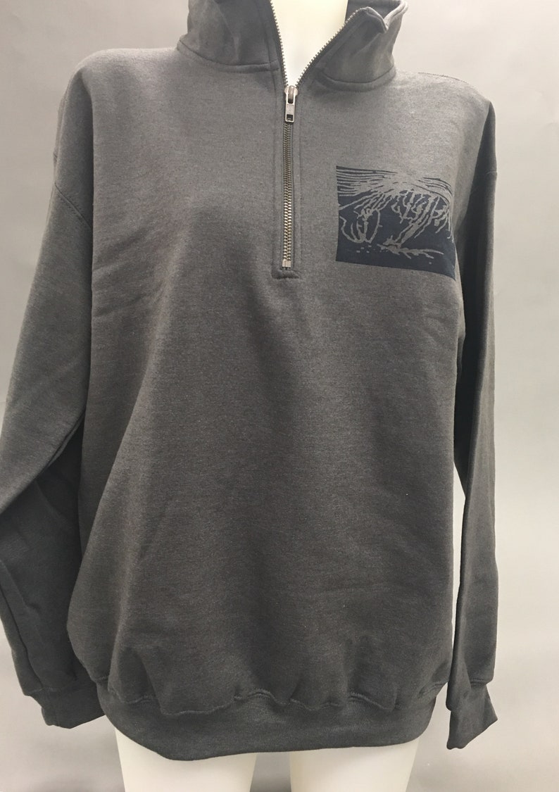 May include: A charcoal gray sweatshirt with a quarter-zip neckline and a small, dark blue square patch on the chest. The patch features a stylized, abstract design. The long-sleeved top is displayed on a mannequin.