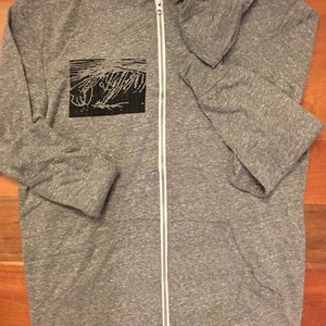 May include: A heather gray zip-up hoodie with a black graphic on the left chest. The hoodie has a white zipper and a hood. The graphic appears to be an abstract design. The hoodie is made of a soft, comfortable material.