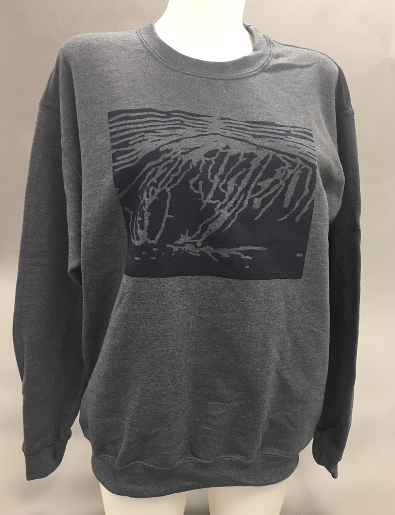 May include: A dark gray sweatshirt with a crew neck and long sleeves. The front features a rectangular graphic in a darker shade of gray, depicting an abstract design. The sweatshirt is displayed on a mannequin.