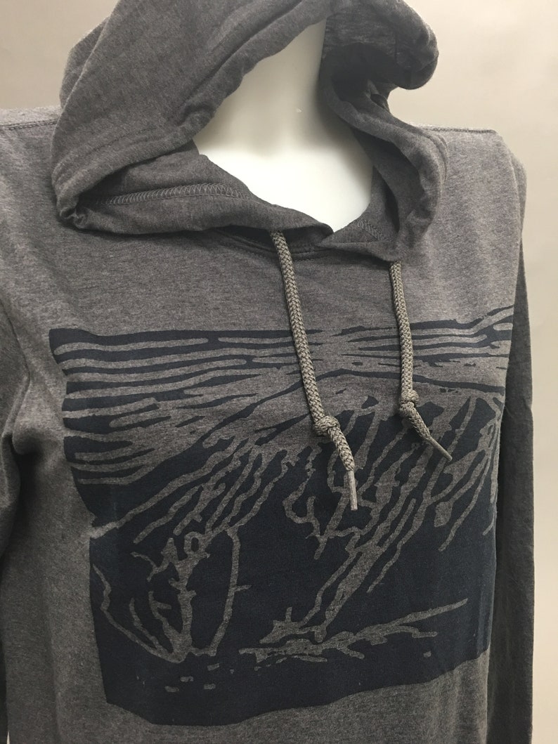 May include: A gray hooded sweatshirt with a dark blue graphic design on the front. The graphic appears to be a stylized landscape or mountain scene. The sweatshirt has a drawstring and a hood.