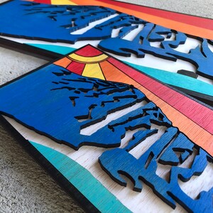 May include: Two wooden wall art pieces featuring a blue mountain range with a layered sunset background. The top piece has a turquoise, orange, and red sunset. The bottom piece has a turquoise, orange, yellow, and red sunset.