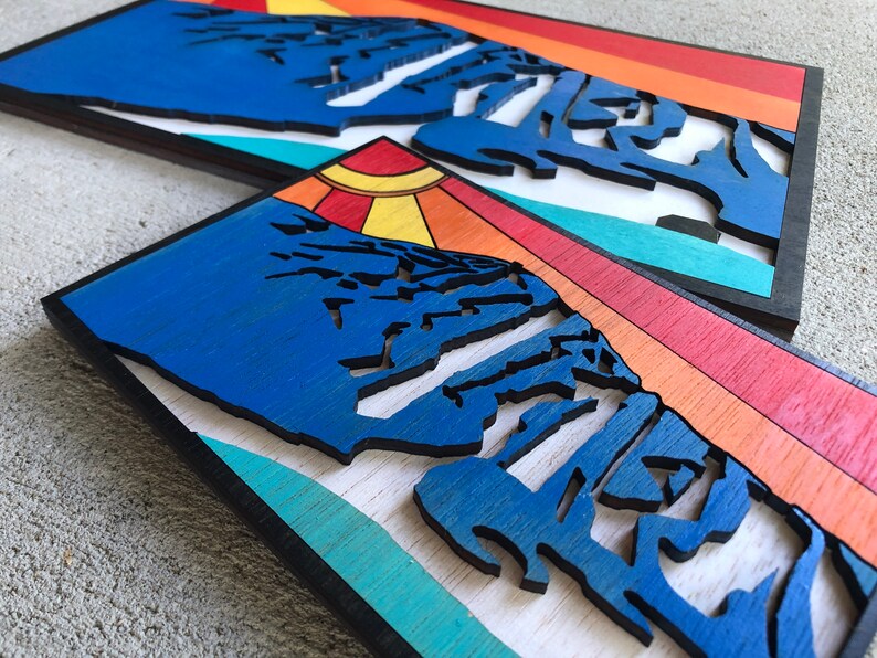 May include: Two wooden wall art pieces featuring a blue mountain range with a layered sunset background. The top piece has a turquoise, orange, and red sunset. The bottom piece has a turquoise, orange, yellow, and red sunset.