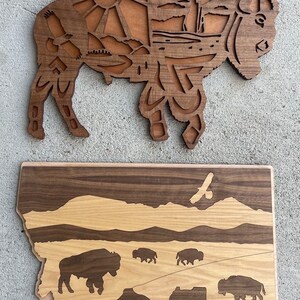 May include: Two wooden wall art pieces depicting the state of Montana. The top piece is a silhouette of a bison with intricate details inside the silhouette. The bottom piece is an outline of the state of Montana with a mountain range, a river, and a herd of bison. Both pieces are made of wood with a natural finish.