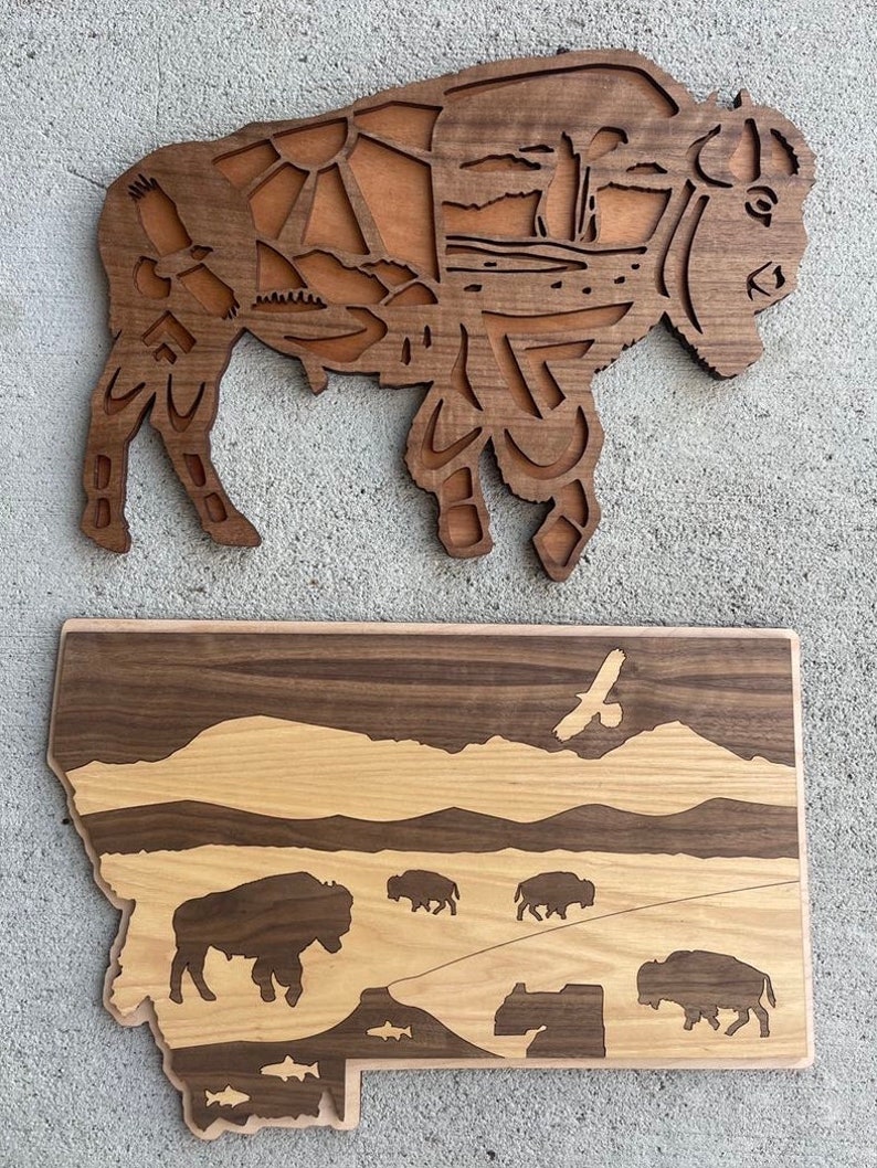May include: Two wooden wall art pieces depicting the state of Montana. The top piece is a silhouette of a bison with intricate details inside the silhouette. The bottom piece is an outline of the state of Montana with a mountain range, a river, and a herd of bison. Both pieces are made of wood with a natural finish.