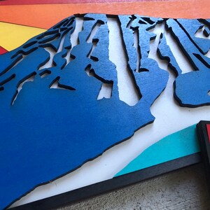 May include: A blue wooden mountain sculpture with a layered, textured design. The mountain is set against a white background with a turquoise and black border.