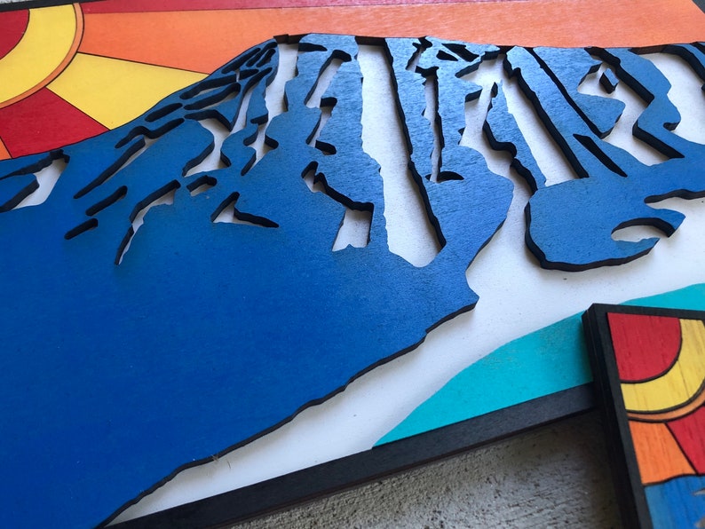 May include: A blue wooden mountain sculpture with a layered, textured design. The mountain is set against a white background with a turquoise and black border.