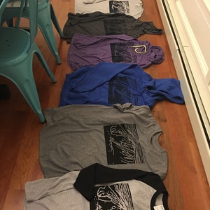 May include: A collection of folded t-shirts and hoodies in various colors, including gray, blue, and purple, are arranged on a wooden floor. Each shirt features a black graphic print of a tree-like design. A black and gray baseball tee is also visible.