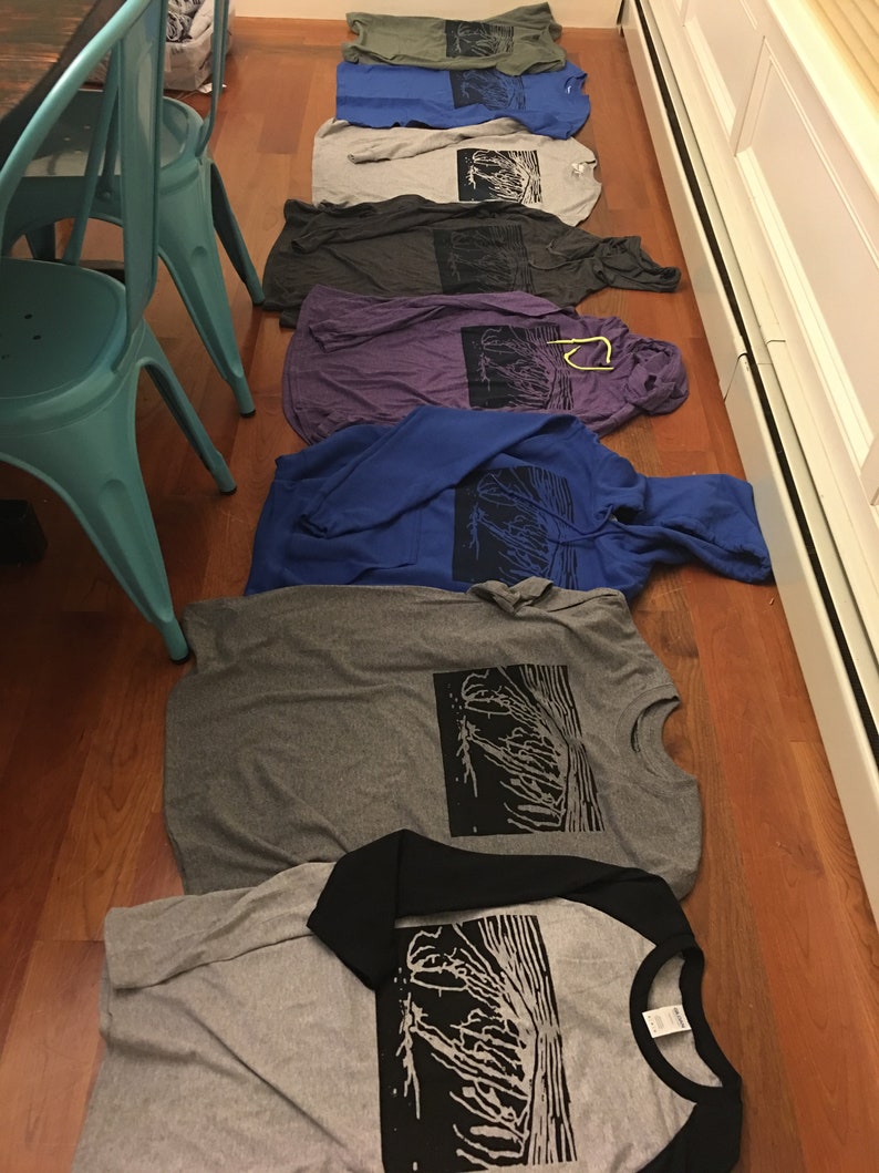 May include: A collection of folded t-shirts and hoodies in various colors, including gray, blue, and purple, are arranged on a wooden floor. Each shirt features a black graphic print of a tree-like design. A black and gray baseball tee is also visible.