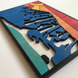 May include: A wooden wall art piece with a blue mountain silhouette against a sunset background. The mountain is layered and textured, with a teal and green base. The sunset is a gradient of orange, yellow, and red.