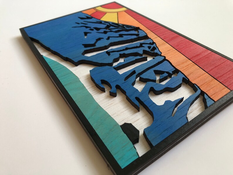 May include: A wooden wall art piece with a blue mountain silhouette against a sunset background. The mountain is layered and textured, with a teal and green base. The sunset is a gradient of orange, yellow, and red.