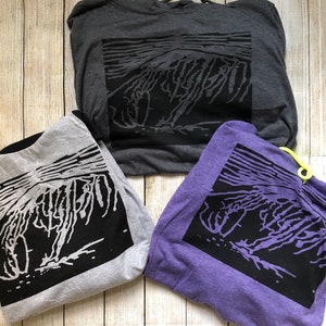 May include: Three hoodies in gray, light gray, and purple, each featuring a black graphic design. The design appears to be an abstract representation of a mountain or landscape. The hoodies are folded and arranged on a light wood-grain surface.