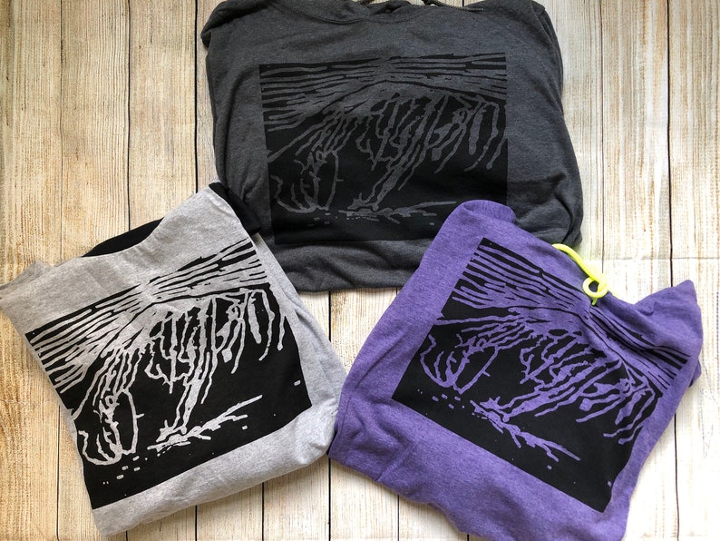 May include: Three hoodies in gray, light gray, and purple, each featuring a black graphic design. The design appears to be an abstract representation of a mountain or landscape. The hoodies are folded and arranged on a light wood-grain surface.