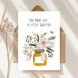 May include: A white greeting card featuring a watercolor illustration of a honey jar filled with pink and green flowers, a small bee, and the text YOU MAKE LIFE A LITTLE SWEETER in black capital letters, centered above the floral arrangement.