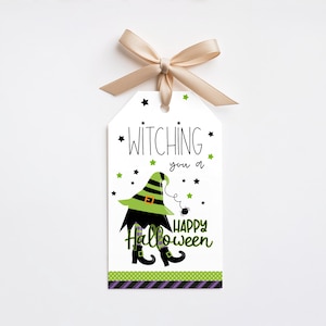 Op de afbeelding: Een rechthoekig wit cadeaulabel met een beige strik aan de bovenkant. Het label bevat de tekst "WITCHING you a HAPPY Halloween" in zwart en groen, met een afbeelding van heksenbenen, een groene hoed en kleine zwarte en groene sterren.