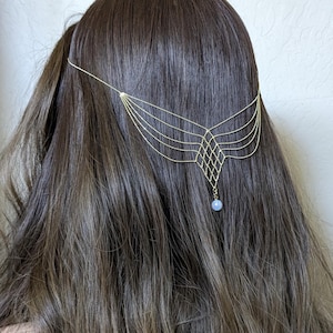 Puede incluir: Un tocado de tono dorado con múltiples cadenas de cuentas delicadas que se extienden sobre el cabello. Las cadenas forman un patrón geométrico de celosía en forma de diamante en el centro, que termina en un pequeño colgante de cuenta azul.