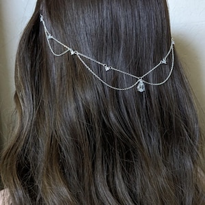 Op de afbeelding: Een zilverkleurig haarketting-accessoire met twee delicate gedrapeerde kettingen. Het ontwerp bevat kleine, heldere, stervormige accenten langs de schakels en een enkele, centrale, druppelvormige heldere kristallen hanger aan de onderste ketting.