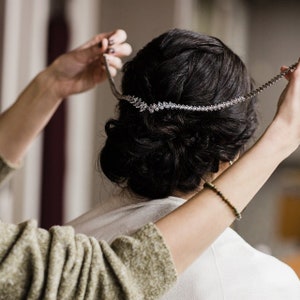 May include: A person putting on a crystal bridal hairpiece. The hairpiece is silver and has a delicate design.