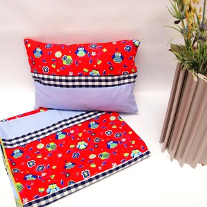 May include: A red pillow and matching blanket set with an owl and floral pattern. The set features a blue and white gingham check border and a light blue center panel. The pillow is rectangular, and the blanket is folded.