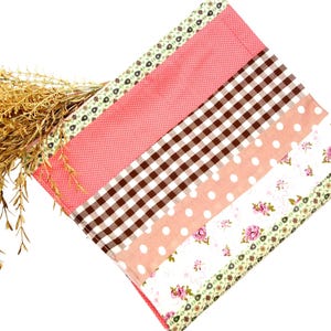 Könnte beinhalten: Ein Patchwork-Stoffartikel mit einer Vielzahl von Mustern, darunter rosa Punkte, braunes Vichy-Karo und Blumendrucke. Der Artikel ist mit einer floralen Bordüre verziert und enthält getrocknete Weizenhalme.