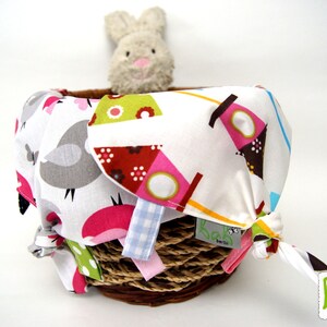 May include: A white fabric basket filled with colorful fabric squares with a bird and geometric patterns. The basket is filled with a stuffed bunny toy. The basket has a brown rope handle and a label that says "bab berlin".