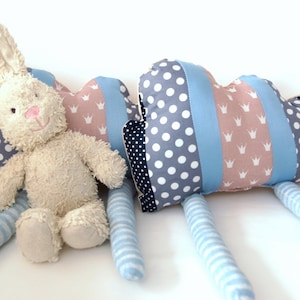 Può includere: Un peluche a forma di coniglietto bianco con un naso rosa e un viso sorridente, accanto a un cuscino a forma di cuore. Il cuscino presenta un motivo a corona rosa e bianca, strisce blu e pois grigi. Il coniglietto ha zampe a righe blu.
