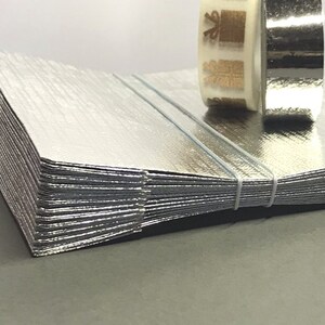 Könnte beinhalten: Ein Stapel silberner Metallfolien mit strukturierter Oberfläche, mit weißer Schnur gebunden. Zwei Rollen Klebeband liegen obenauf; eine ist silberfarben, die andere ist transparent mit goldfarbenen Designs. Der Hintergrund ist schlicht weiß.
