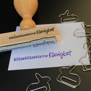 Könnte beinhalten: Ein Holzstempel mit dem Text "klitzeklitzekleine Kleinigkeit" wird gezeigt, zusammen mit einem lila Tintenabdruck desselben Textes auf weißem Papier. Mehrere silberne hasenförmige Büroklammern sind verstreut.
