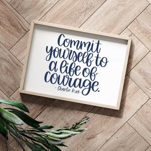 May include: A framed print with the text "Commit yourself to a life of courage" in navy blue script. The print is in a light wood frame, resting on a wooden surface with a sprig of green leaves.