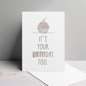 May include: A white birthday card with a line drawing of a cupcake topped with a cherry. The text on the card reads "IT'S YOUR BIRTHDAY TOO." A white envelope is visible below the card. The card is on a light wood surface.