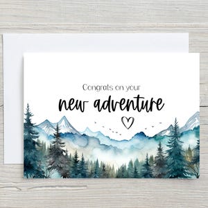 May include: A greeting card with a watercolor landscape of mountains and trees in shades of blue and green. The card reads "Congrats on your new adventure" with a heart symbol.