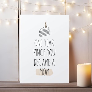 May include: A white card with the text "ONE YEAR SINCE YOU BECAME A MOM" and a cake illustration. A lit candle and string lights are in the background. The card is on a wooden surface.