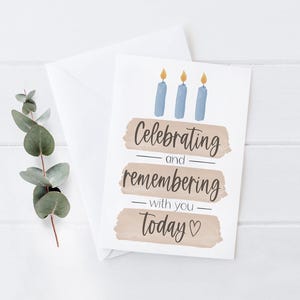 May include: A white greeting card with the text "Celebrating and remembering with you today" and three blue candles with yellow flames. The card is accompanied by a white envelope and a sprig of green leaves.