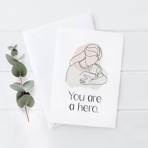 Pode incluir: Um cartão branco com um desenho de linha de um pai segurando uma criança, com um fundo rosa e cinza suave. O texto "You are a hero" é impresso em preto. Um envelope e um ramo de vegetação estão no fundo.