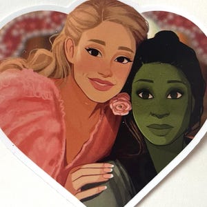 May include: A heart-shaped sticker featuring two women. One woman has blonde hair and wears a pink dress, the other has green skin and black hair. A pink rose is near the blonde woman's face. The sticker has a white border.