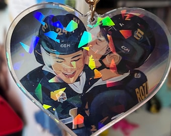 Könnte beinhalten: Ein herzförmiger Schlüsselanhänger mit einem holografischen Bild von zwei Hockeyspielern in schwarzen und marineblauen Uniformen. Ein Spieler trägt die Nummer 24 und der andere den Namen "ROZ" auf seinem Trikot. Der Schlüsselanhänger hat einen goldfarbenen Verschluss.
