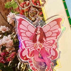 May include: A pink and red butterfly-shaped keychain with a silver-colored clasp. The keychain features a drawing of a woman with red hair and a pink outfit. The butterfly wings have a holographic effect.