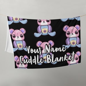 May include: A black cuddle blanket with a repeating pattern of pink and purple teddy bears with stitched details. The text "Your Name Cuddle Blanket" is printed in white on the blanket.