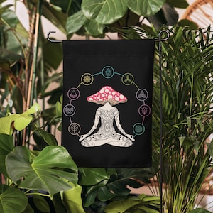 May include: Black garden flag featuring a mushroom-shaped figure in a meditative pose. The mushroom has a pink cap with white spots. A circular design with colorful symbols surrounds the figure. The flag is displayed outdoors among green plants.