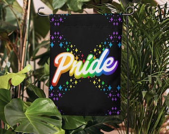 May include: A black garden flag with the word "Pride" in rainbow colors. The flag has a pattern of colorful diamond shapes. The flag is displayed outdoors among green plants.