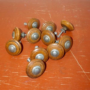 Puede incluir: Un conjunto de tiradores de madera para armarios con detalles metálicos plateados. Los tiradores son redondos con una parte superior plana y un tornillo para fijarlos. La madera tiene un tono marrón cálido, y los tiradores están sobre una superficie marrón rojiza.