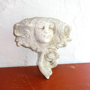 May include: A white plaster wall sculpture of a woman's face with closed eyes, surrounded by flowing hair and floral details. The sculpture is mounted on a red surface.