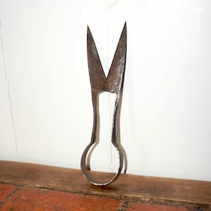 May include: Vintage metal scissors with a loop handle and pointed blades. The scissors are made of a dark, aged metal, and are set against a white wall and a wooden surface. The blades are open, ready for use.