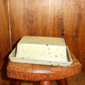 Könnte beinhalten: Eine hellgrüne Keramik-Butterdose mit Deckel, gesprenkelt mit dunkelbraunen Punkten. Die Dose steht auf einem kleinen, dreibeinigen Holzschemel. Der Hintergrund ist eine Holzwand.
