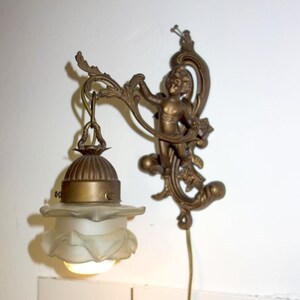 May include: Antique brass wall sconce featuring a cherub figure and floral accents. The sconce has a frosted glass shade and a decorative design, adding a vintage touch to any space.